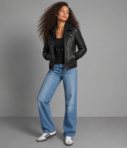 A model wearing a black leather moto jacket, black top, wide-leg blue jeans, and white sneakers, standing against a solid gray studio background.