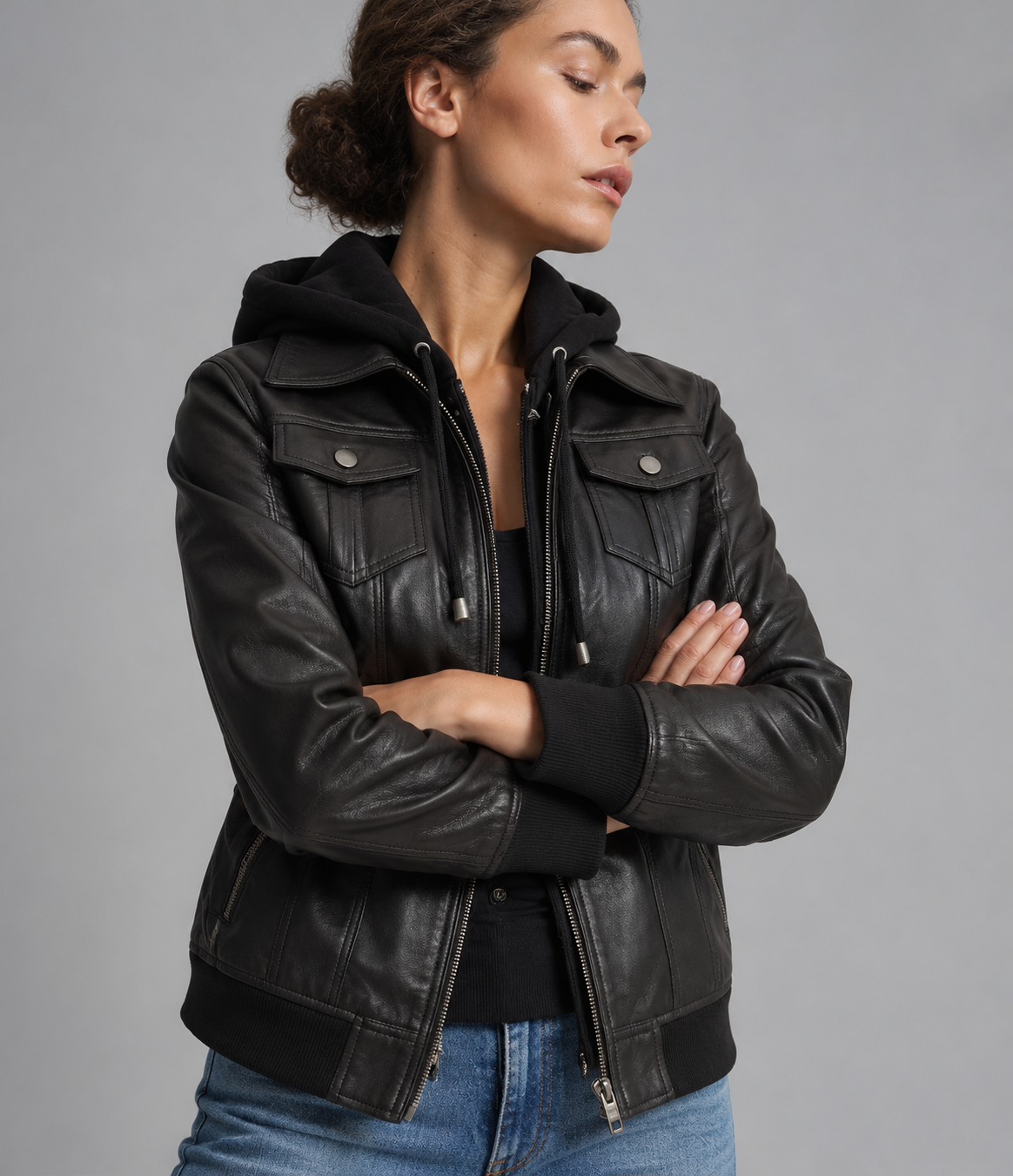 A woman wearing a black leather moto jacket with a built-in black hood, styled over a black top and blue jeans, photographed against a solid gray studio background.