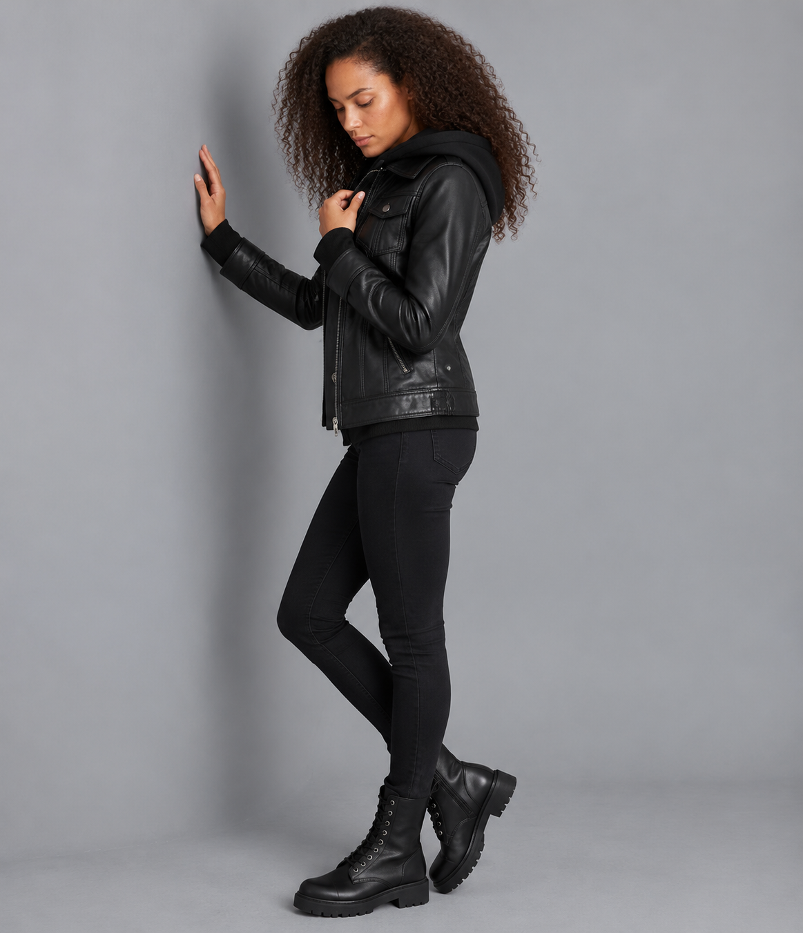 A woman modeling a black faux-leather moto jacket with a hood, paired with black skinny pants and black combat boots, posing against a solid gray studio background.