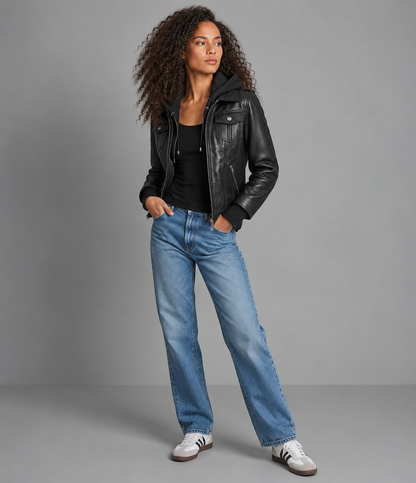A model with curly hair stands in a studio wearing a black leather moto jacket, a black top, blue jeans, and white sneakers with black stripes, against a solid gray background.