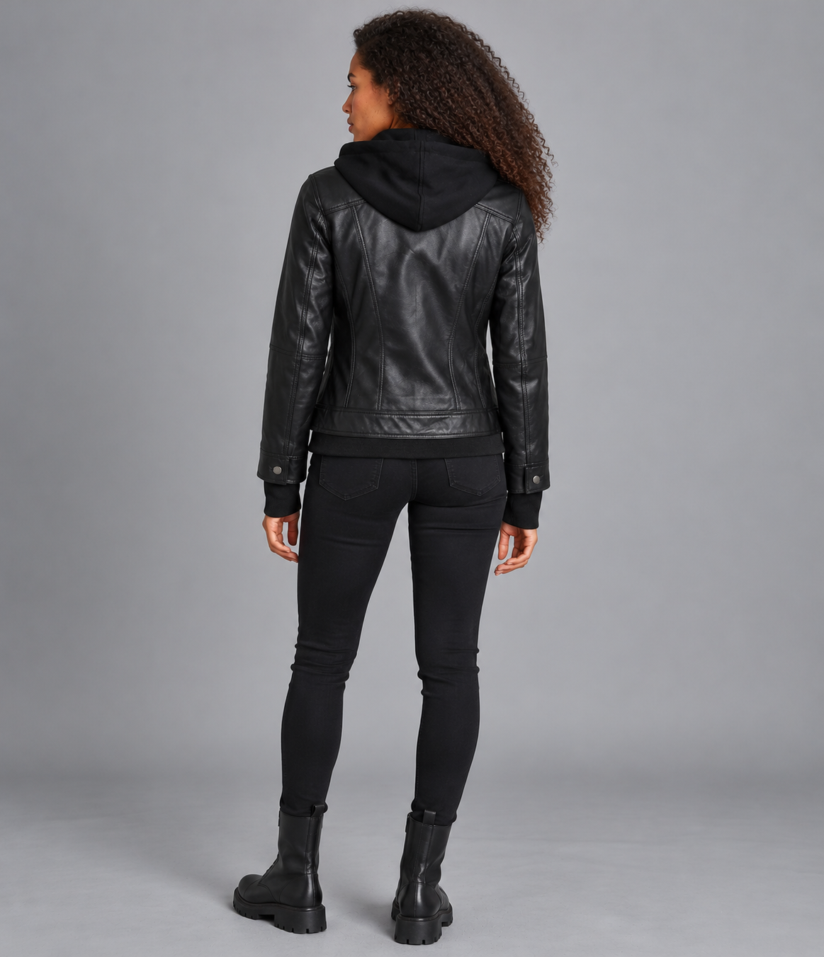 A woman shown from the back wearing a black leather hooded jacket, black skinny jeans, and black boots against a solid gray studio background.