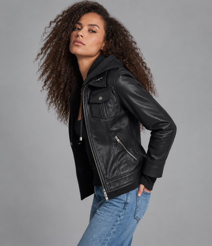 A woman with long curly brown hair models a black leather moto jacket with a hood, worn over a black top and paired with blue jeans, against a solid gray studio background.